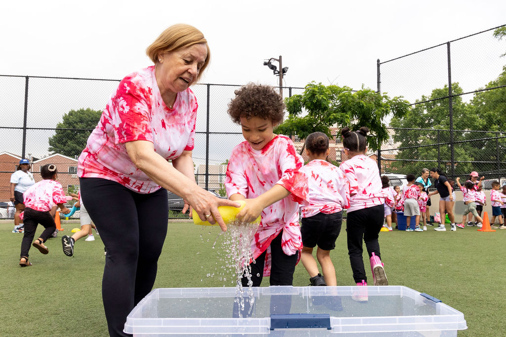 Adult and child in matching pink tie-dye shirts working together on a water activity on a sports field