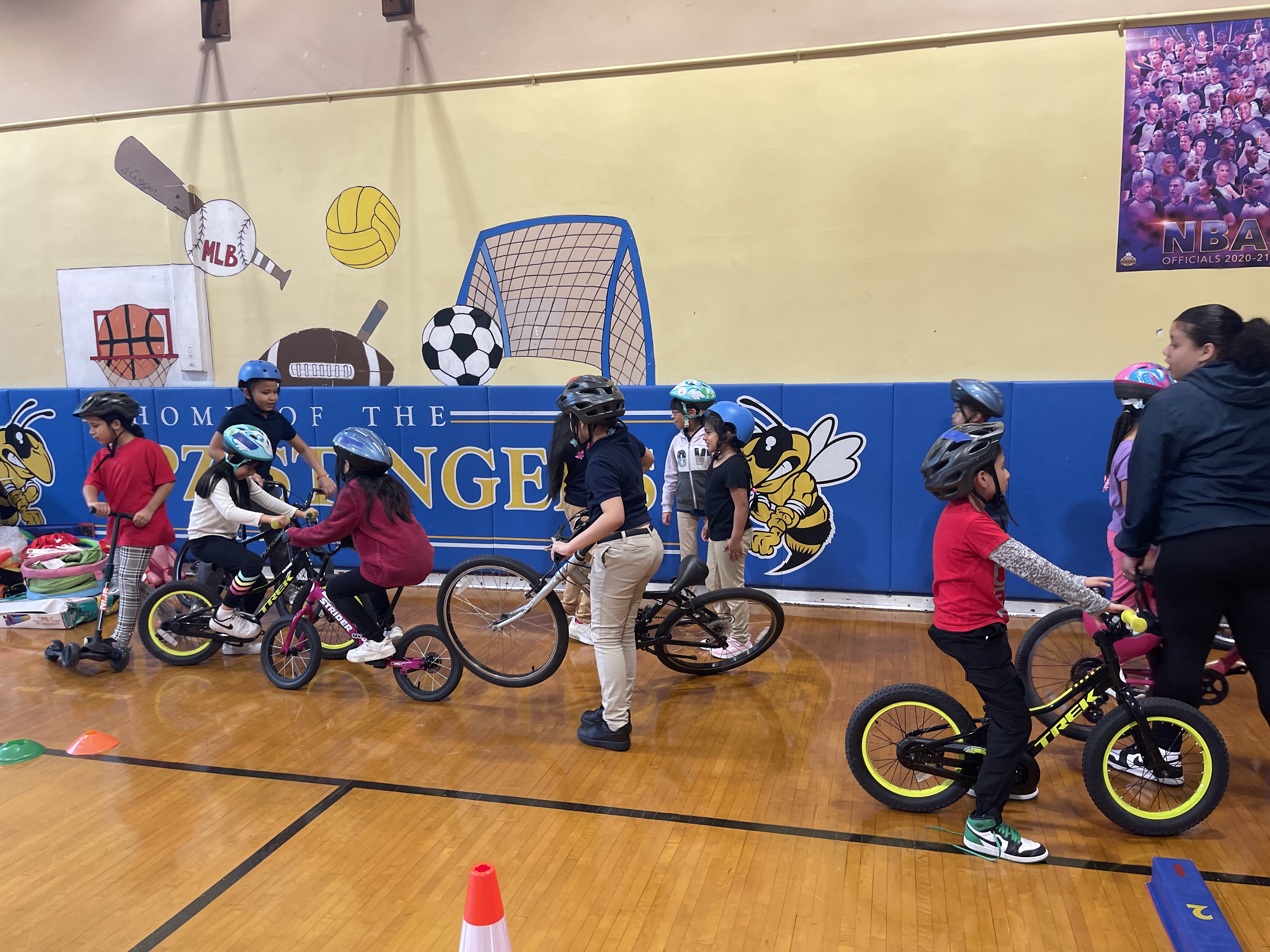 Children learning to ride bicycles in a school gymnasium with 'Home of the Hornets' banner