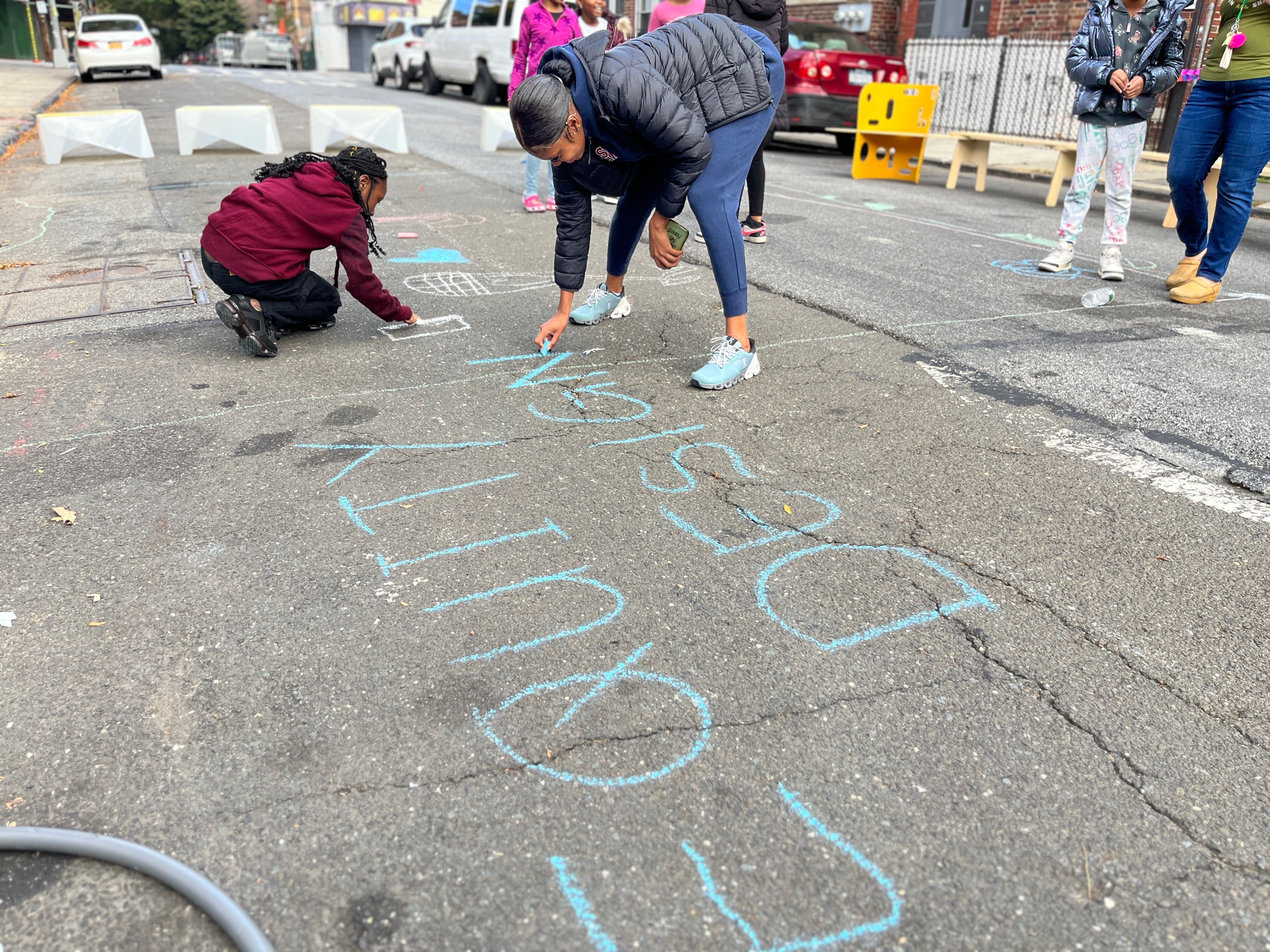 Community members drawing with chalk on a street, writing 'WELCOME' as part of a physical activity program